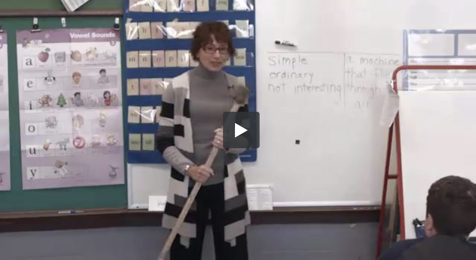 A teacher stands holding a large pointer stick in front of a whiteboard in a classroom. The whiteboard has text that reads Simple, ordinary, not interesting. Classroom posters and charts related to vowel sounds are visible on the wall beside her. Students are sitting at desks.